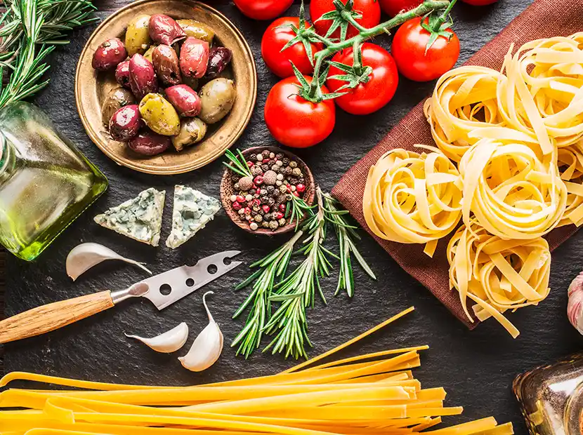 Tisch mit Zutaten wie Pasta, Oliven, Tomaten, Rosmarin, Pfeffer und Gorgonzola.